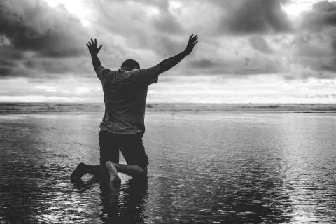Man on his knees on the shore of a lake under a cloudy sky in black and white by Forgiven Photography | Lightstock | Used by Permission